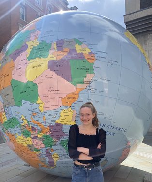 UW/H-Alumna Anna Stricker (Foto: Privat) Eine junge Frau steht mit verschränkten Armen vor einer riesigen Skulptur einer Weltkugel und lächelt in die Kamera.