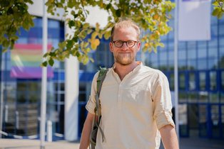 UW/H-Alumnus Christian hat PPE studiert und ist in die Politik gegangen. (Foto: UW/H) Porträt eines Mannes vor dem Universitätsgebäude. Über der Schulter trägt er einen Rucksack.
