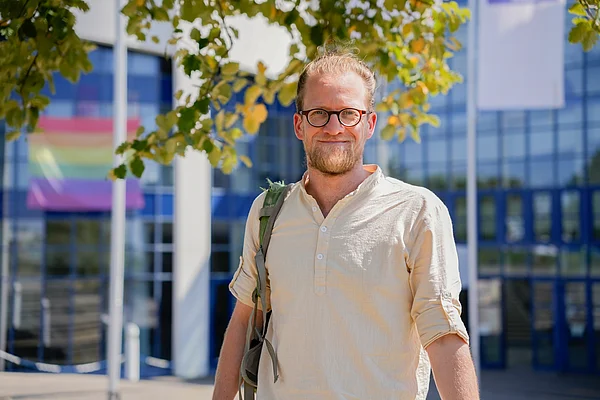 Porträt eines Mannes vor dem Universitätsgebäude. Über der Schulter trägt er einen Rucksack.