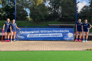 Vier Hockey-Spielerinnen flankieren ein blaues Werbebanner der Uni Witten/Herdecke am Spielfeldrand eines Hockeyfeldes.