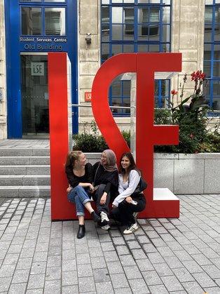 UW/H-Alumna Anna Stricker vor der London School of Economics (Foto: privat) Drei junge Frauen sitzen auf einer Skulptur aus Buchstaben.
