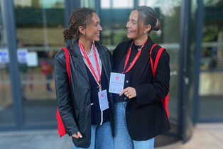 Foto von Clara und Camille vor einer Glastür. Beide lächeln sich an.