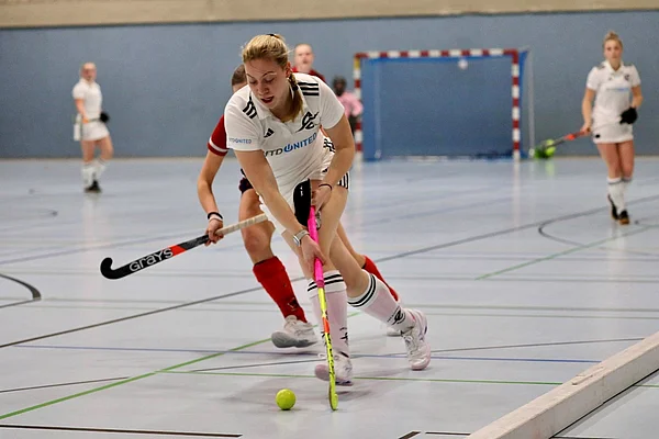 Eine Hockey-Spielerin im Zweikampf um den Ball. Das Spiel findet in einer Sporthalle statt.