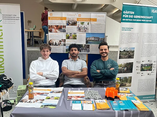 Sami Barkal sitzt mit zwei Kommilitonen an einem Infotisch. Hinter ihm stehen eine Pinnwand und ein Banner mit Informationen zu Stipendien.