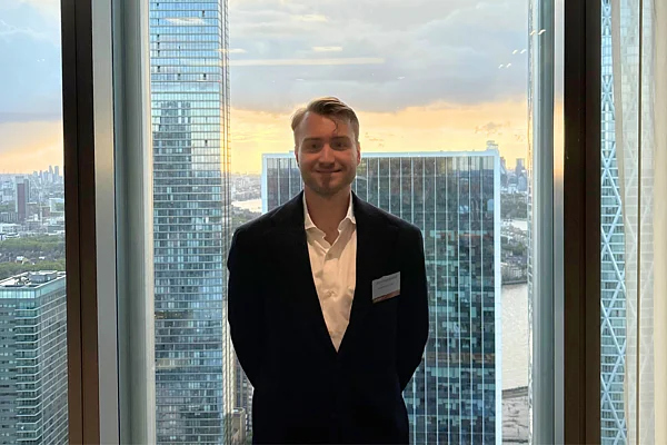 Ein Mann im Anzug steht vor einer Fensterfront mit Hochhäusern im Hintergrund.