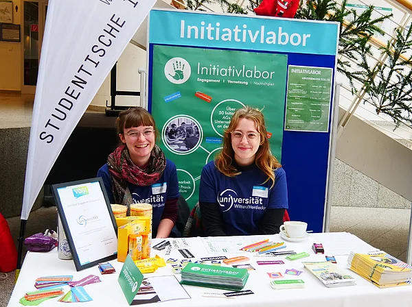 Emmi Mentzel und eine Kommilitonin sitzen an einem Infostand. Im Hintergrund sind eine Beachflag und eine Posterwand mit der Aufschrift "Initiativlabor".