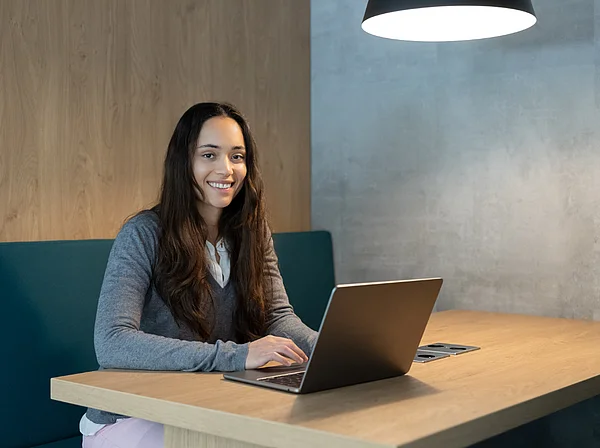 Eine Studentin sitzt mit ihrem Laptop an einem Tisch und lächelt in die Kamera.