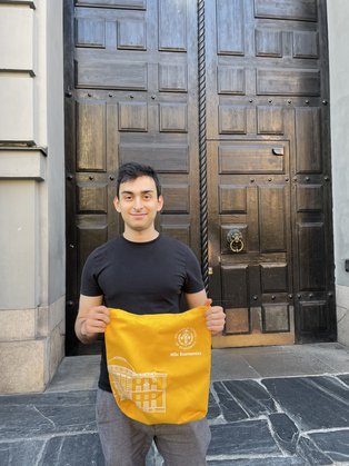 Alumnus Amir Rahimi vor seiner neuen Uni in Stockholm (Foto: privat) Amir Rahimi steht vor einer großen braunen Tür und hält einen orangenen Beutel in die Kamera.
