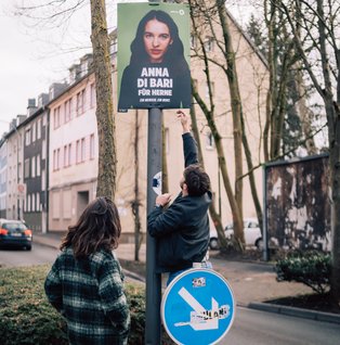 Zwei Personen hängen ein Wahlkampf der Bundestagskandidatin an einem Laternenpfahl auf.