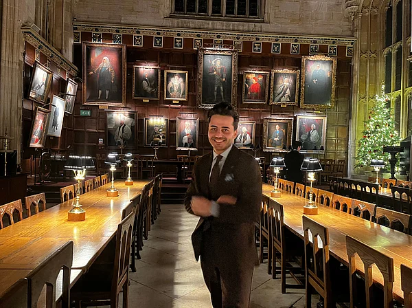 Sami Barkal steht in einem historischem Saal der Oxford University. Hinter ihm hängen Porträts an der Wand. In der hinteren rechten Ecke steht ein beleuchteter Weihnachtsbaum.