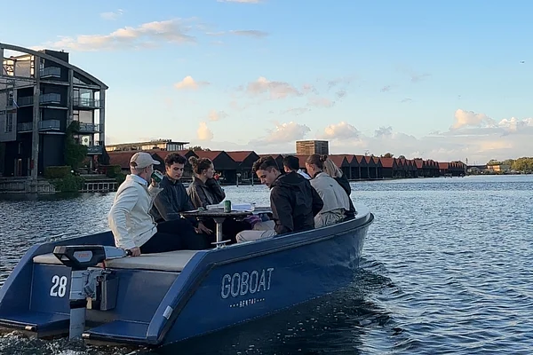 Eine Gruppe von Menschen sitzt in einem Boot auf dem Wasser. Im Hintergrund sind Gebäude.